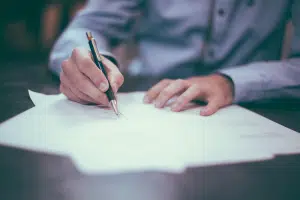 A man in a blue button down shirt signing papers on a wood table with a pen. 